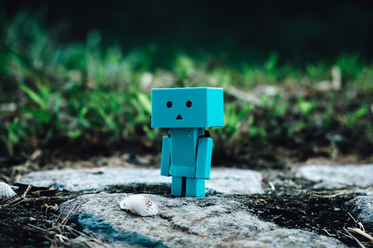 gallery-01 Cute blue toy robot standing on a rocky path surrounded by greenery.